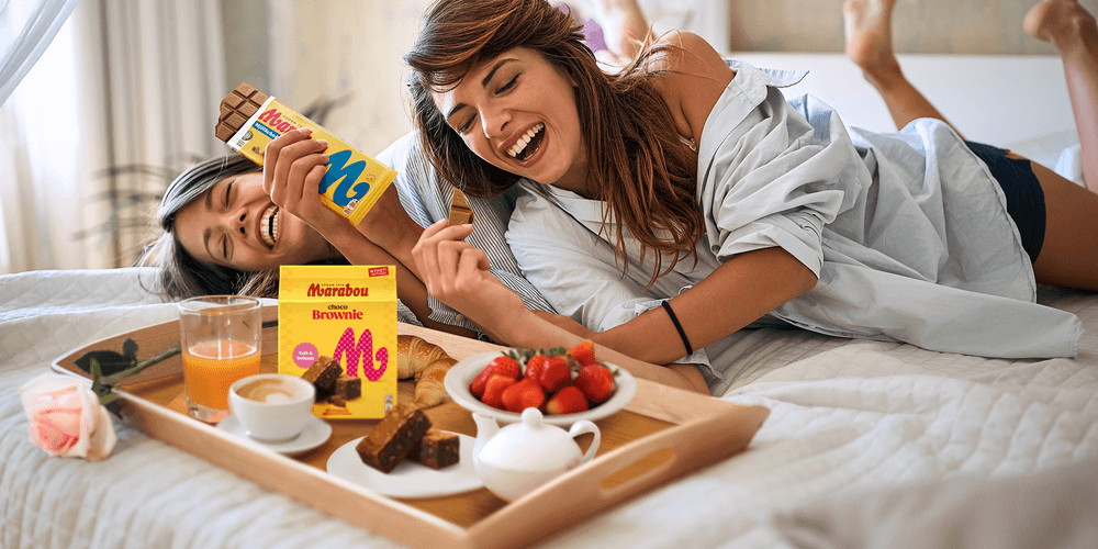 Två kvinnor skrattar på en säng med en frukostbricka som innehåller chokladkakor, juice och jordgubbar.